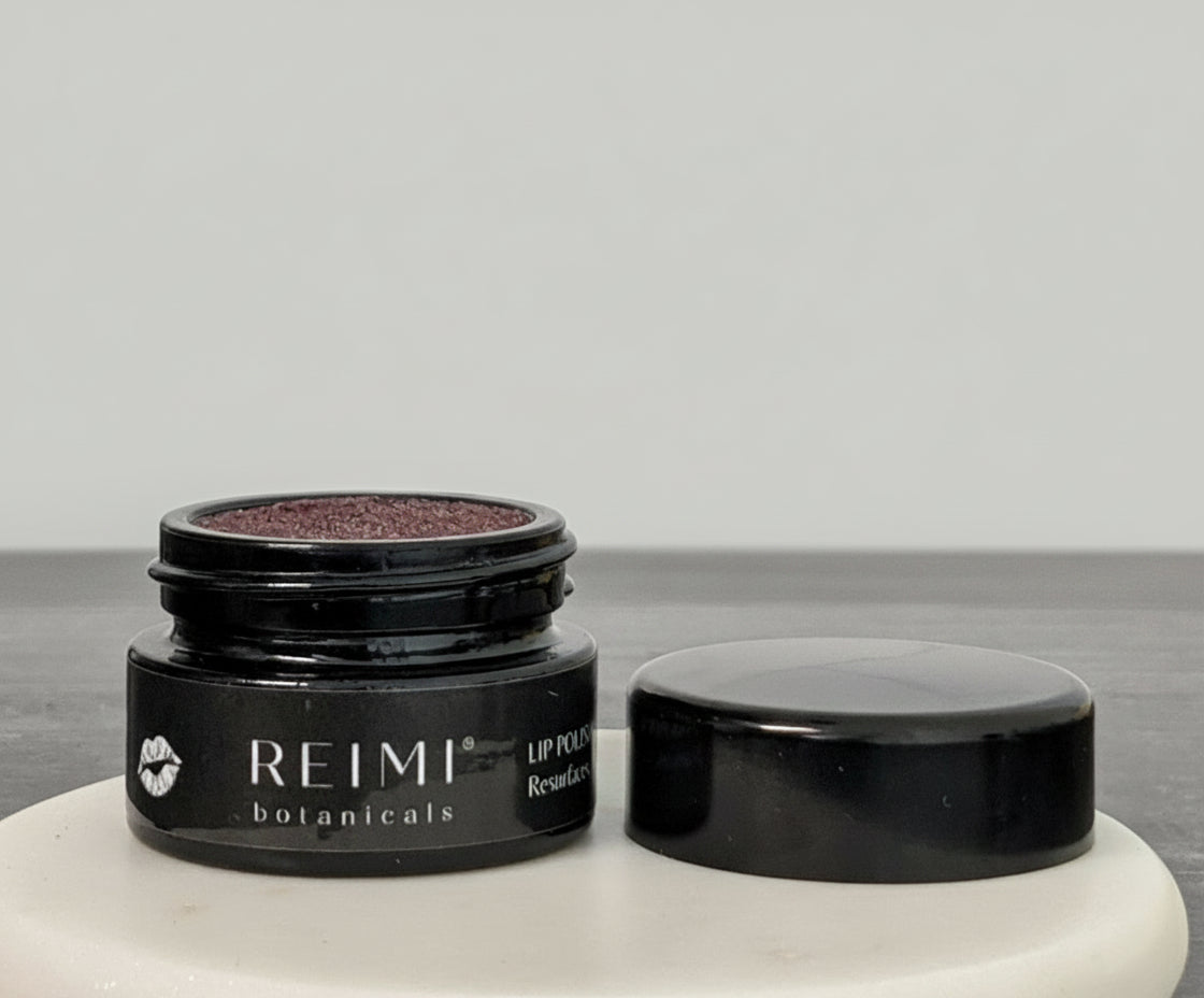 A jar of REIMI Botanicals Lip Polishing Balm with Hibiscus + Black Raspberry. The product is in a black container with a label and the brand name is visible.