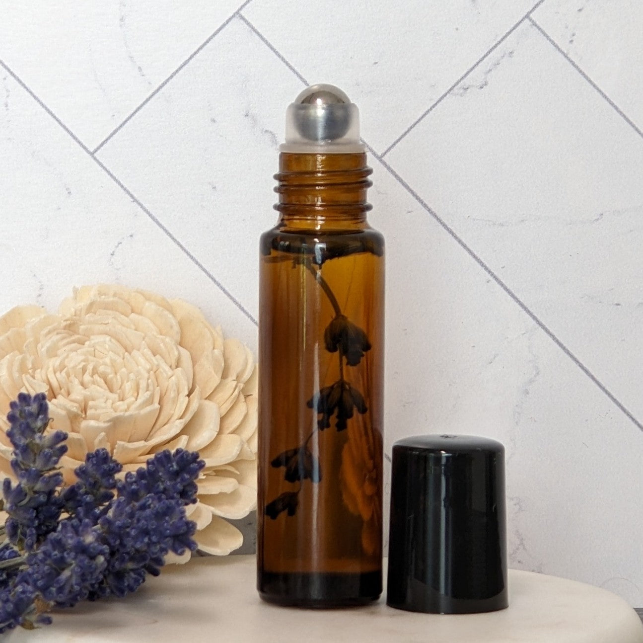 Amber galss roll-on bottle of oil with lavender sprig inside, wite background on marble stand with lavender flowers and white flower