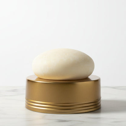 Round white lotion bar on a gold cylindrical container on a white marble countertop