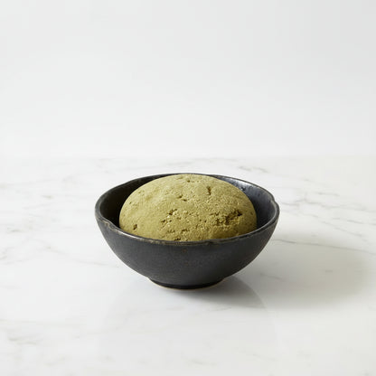 A round, textured olive-green stone or clay object resting inside a dark, scalloped ceramic bowl on a brown kraft paper background.