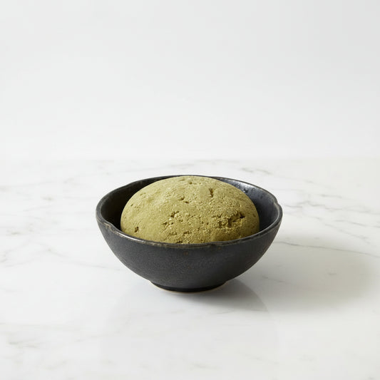 A round, textured olive-green stone or clay object resting inside a dark, scalloped ceramic bowl on a brown kraft paper background.