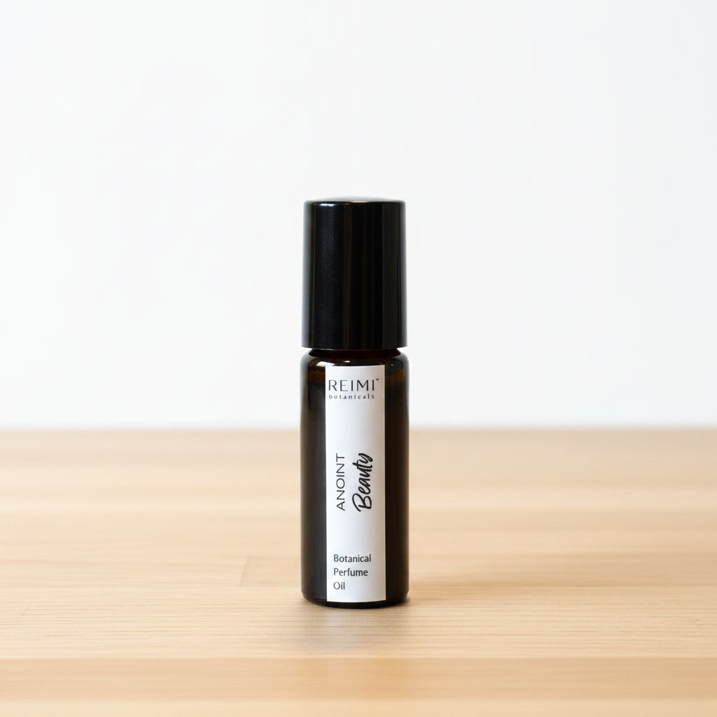 A small black roll-on bottle with a white label reading 'ANOINT  Beauty' and stands on a textured beige surface. The background is a soft-focus indoor setting with neutral tones and partially visible items behind it.