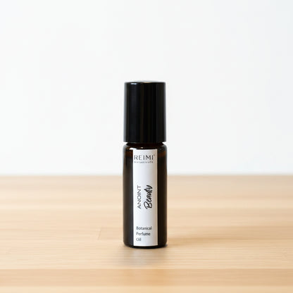 A small black roll-on bottle with a white label reading 'ANOINT  Beauty' and stands on a textured beige surface. The background is a soft-focus indoor setting with neutral tones and partially visible items behind it.