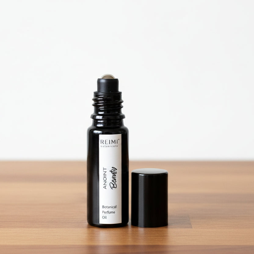 A small black glass roller bottle labeled 'REIMI Botanicals ANOINT Beauty Botanical Perfume Oil' stands next to its black cap on a textured beige fabric surface against a gray fabric backdrop.