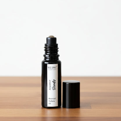 A small black glass roller bottle labeled 'REIMI Botanicals ANOINT Beauty Botanical Perfume Oil' stands next to its black cap on a textured beige fabric surface against a gray fabric backdrop.