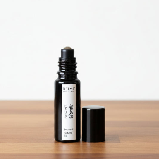 A small black glass roller bottle labeled 'REIMI Botanicals ANOINT Beauty Botanical Perfume Oil' stands next to its black cap on a textured beige fabric surface against a gray fabric backdrop.