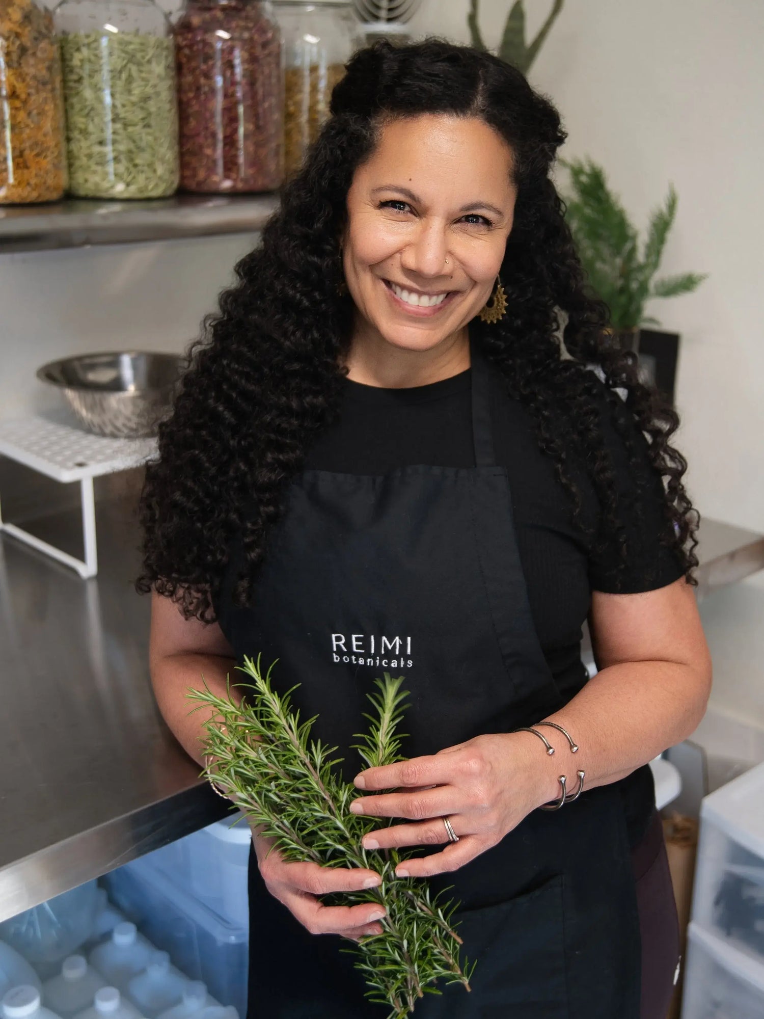 REIMI Botaniclas founder holding a bundle of rosemary in a kitchen setting, wearing a black apron with 'REIMI' branding.